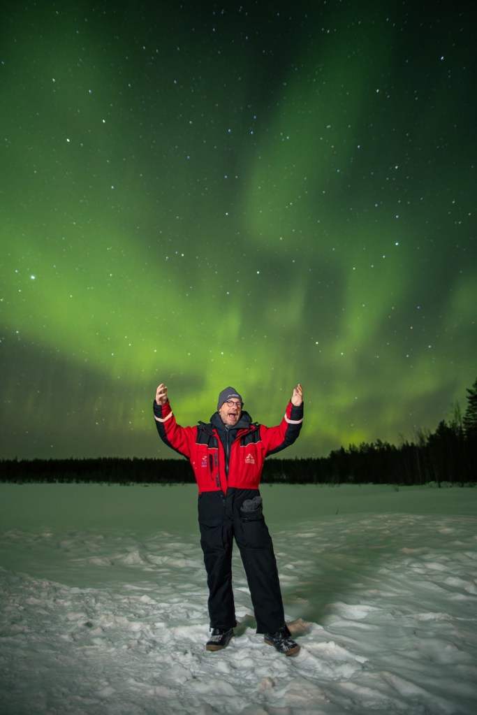 A person in winter clothing stands in a snowy landscape under a vibrant display of the Northern Lights, with green and blue hues illuminating the night sky.