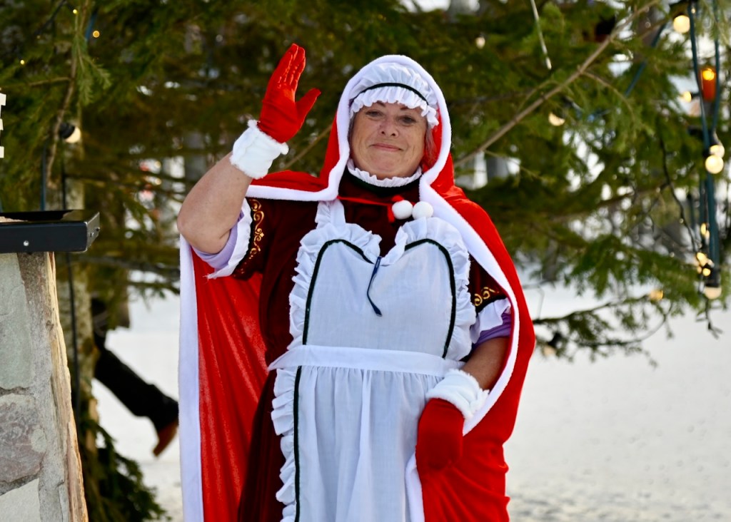 A woman dressed in a festive red and white costume, waving happily, with a backdrop of green trees adorned with lights.