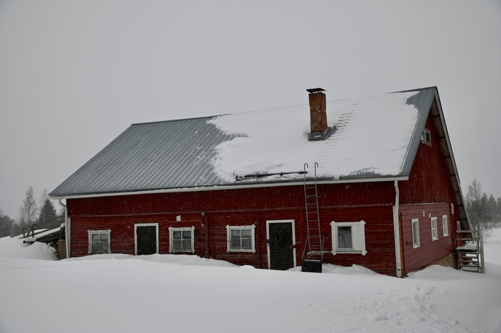 A traditional red wooden house with a sloped roof, covered in snow, featuring a chimney and a ladder leaning against the side, set in a winter landscape.