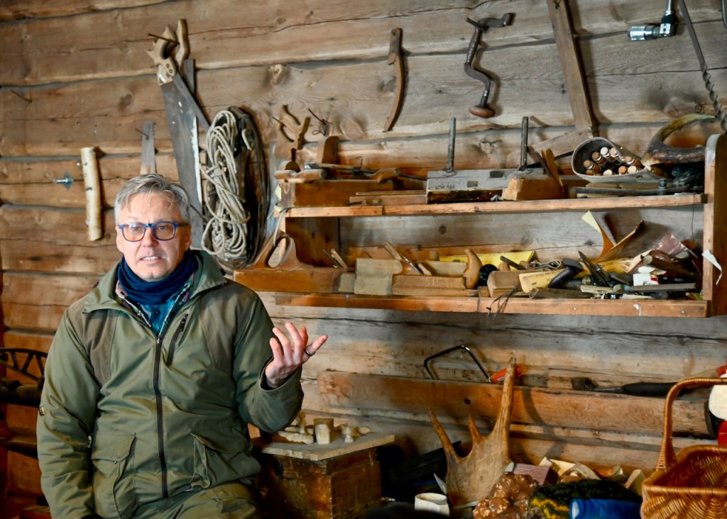 A man seated in a rustic wooden workshop, gesturing while surrounded by various handmade tools and equipment on shelves in the background.