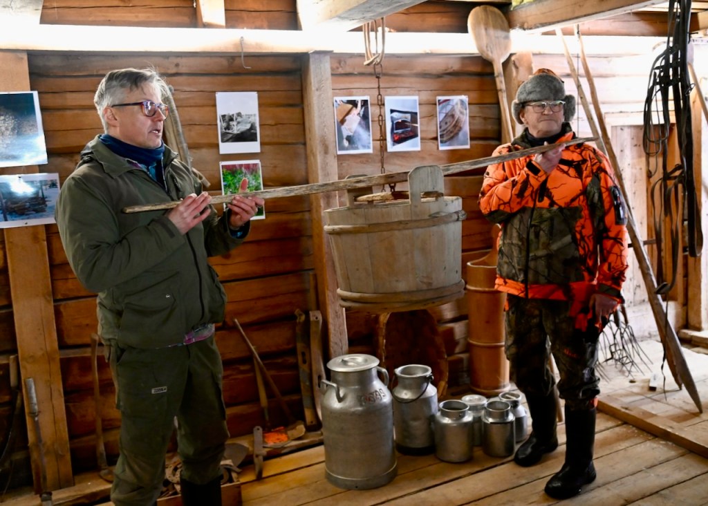 Two men demonstrate traditional wooden tools in a rustic wooden building, surrounded by various milk containers and photographs depicting local culture.