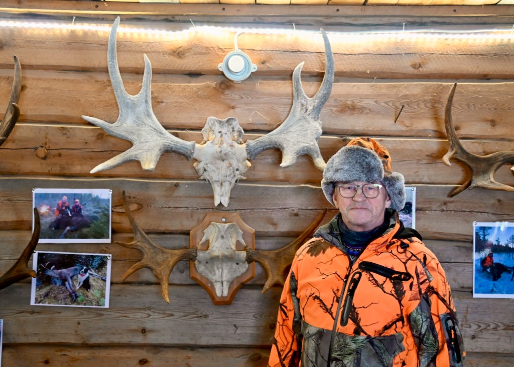 A local man wearing a camouflage jacket and fur hat stands in front of a wooden wall decorated with moose antlers and photographs of hunting activities.