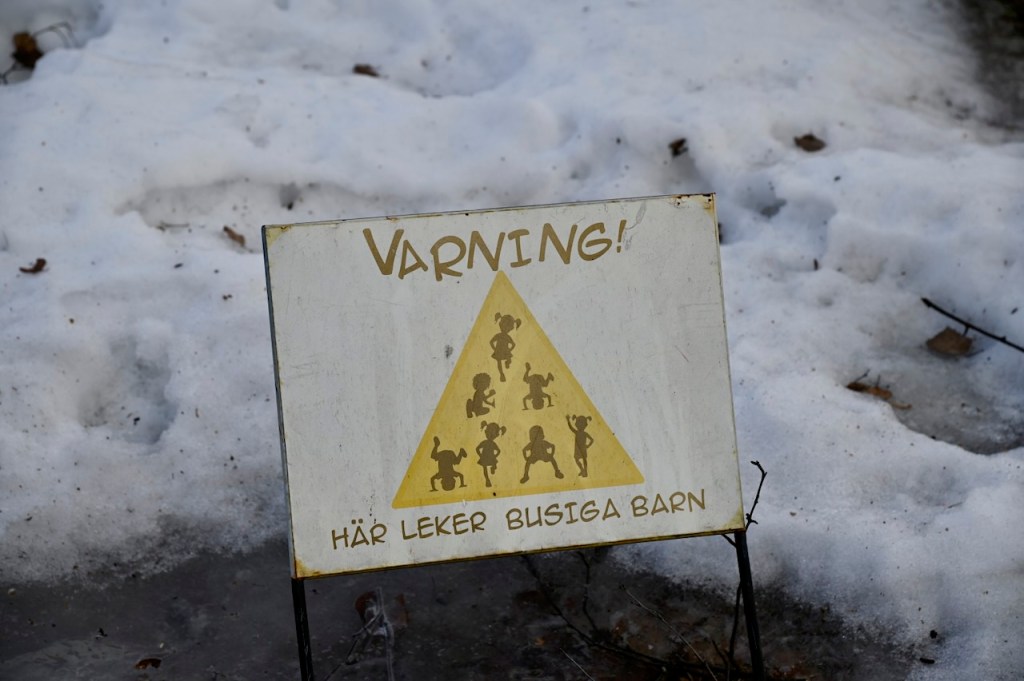 A warning sign in Swedish with playful icons indicating children are playing nearby, set against a snowy background.