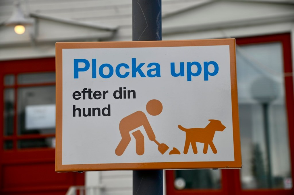 A sign in Swedish instructing dog owners to clean up after their pets, featuring an illustration of a person picking up dog waste.