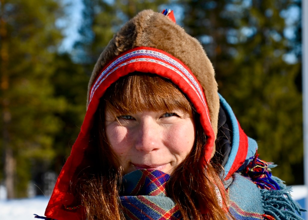 A young woman wearing traditional Sami clothing with a fur hat and colorful scarf, smiling against a snowy forest background.