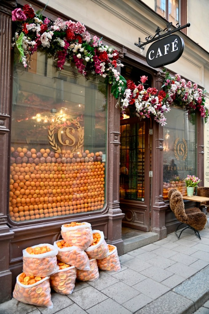 A charming café with a floral display above the entrance, featuring large glass windows filled with oranges and several bags of oranges stacked on the sidewalk.