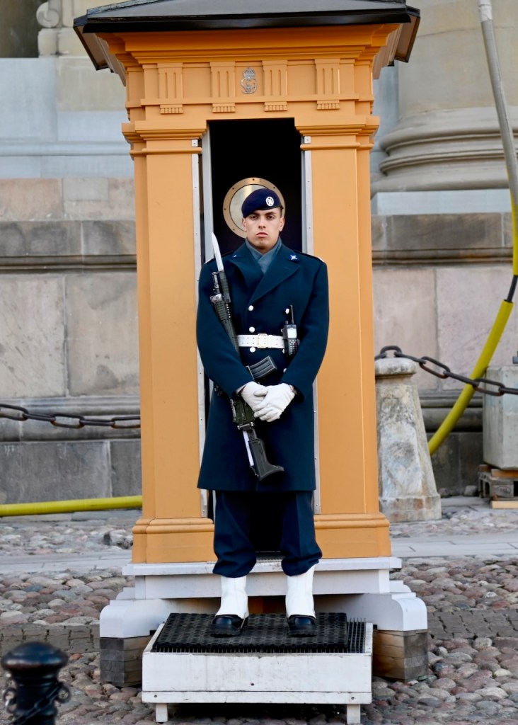 A guard in a military uniform stands at attention in front of a small, ornate yellow guardhouse. The guard is holding a rifle and wearing a hat with insignia.