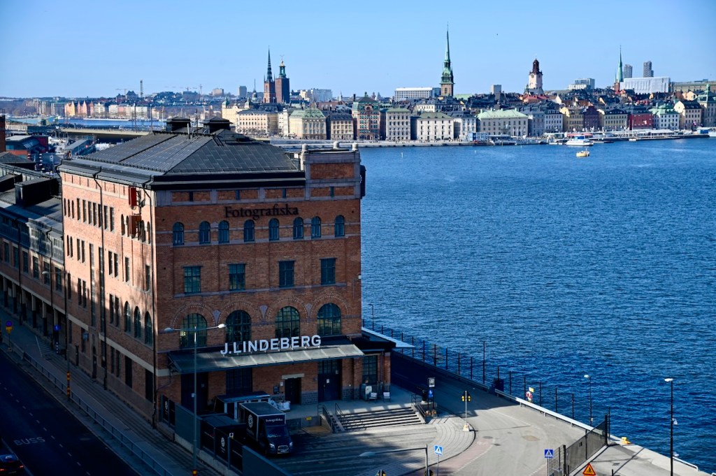 A panoramic view of Stockholm's waterfront, showcasing a historic brick building labeled 'Fotografiska' in the foreground, with the city's skyline and various architectural styles in the background.
