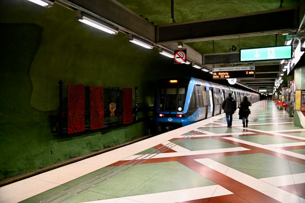 Interior of a Stockholm metro station featuring artistic walls, with a blue train at the platform and two people walking along the tiled floor.