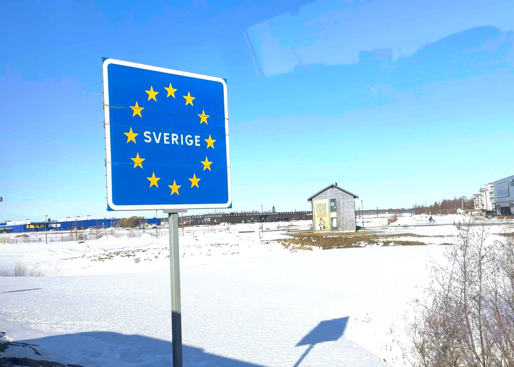 A sign indicating the border crossing into Sweden, featuring the word 'SVERIGE' and surrounded by the European Union stars. The background shows a snowy landscape with a small building and an Ikea store in the distance.