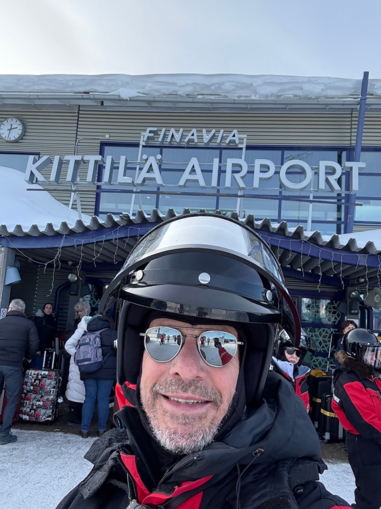 A man wearing a helmet and snowmobiling gear stands in front of Kittilä Airport, with a sign reading 'KITTILÄ AIRPORT' visible. Snow is on the ground and people with luggage are seen in the background.