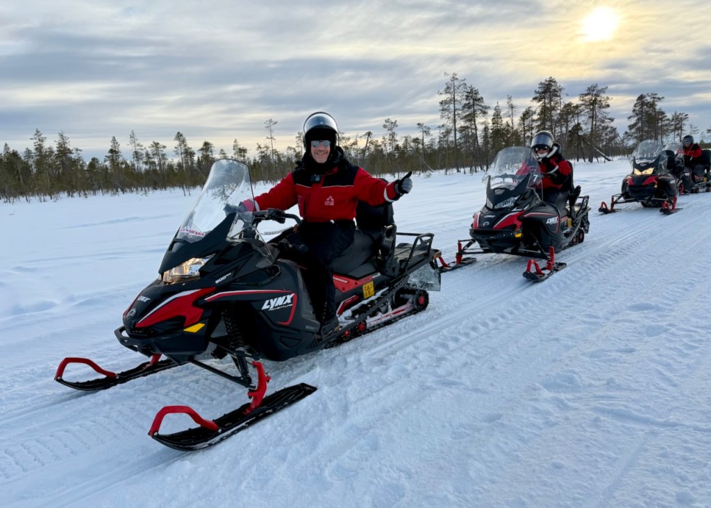 A group of snowmobilers, with one person giving a thumbs up, riding on a snowy landscape in Finnish Lapland.