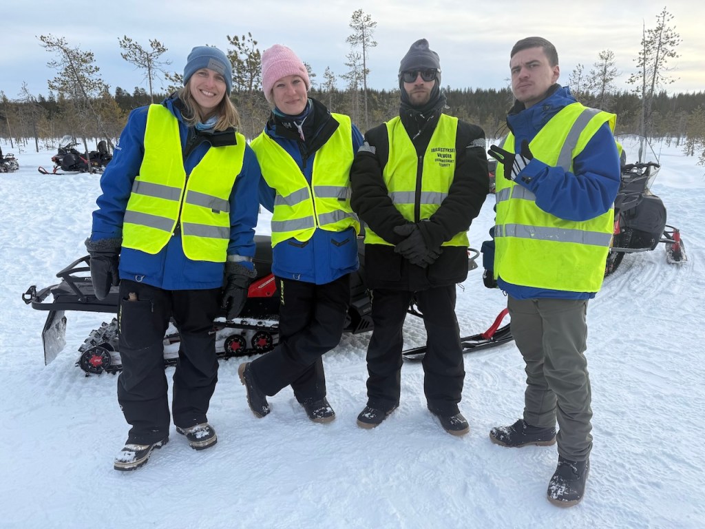 Four snowmobile tour guides in bright yellow vests pose together in a snowy landscape, with snowmobiles parked behind them.