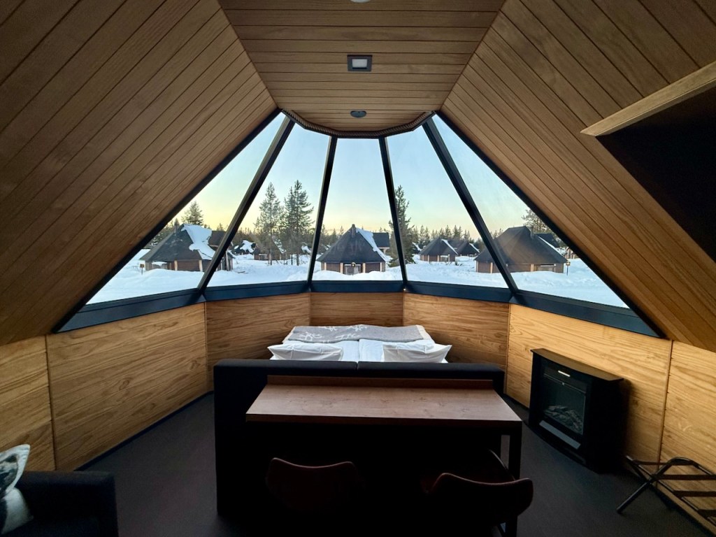 Interior view of an 'Aurora Cabin' in Finnish Lapland, featuring a wooden ceiling, large glass windows offering a snowy landscape view, and a simple seating area with a table.