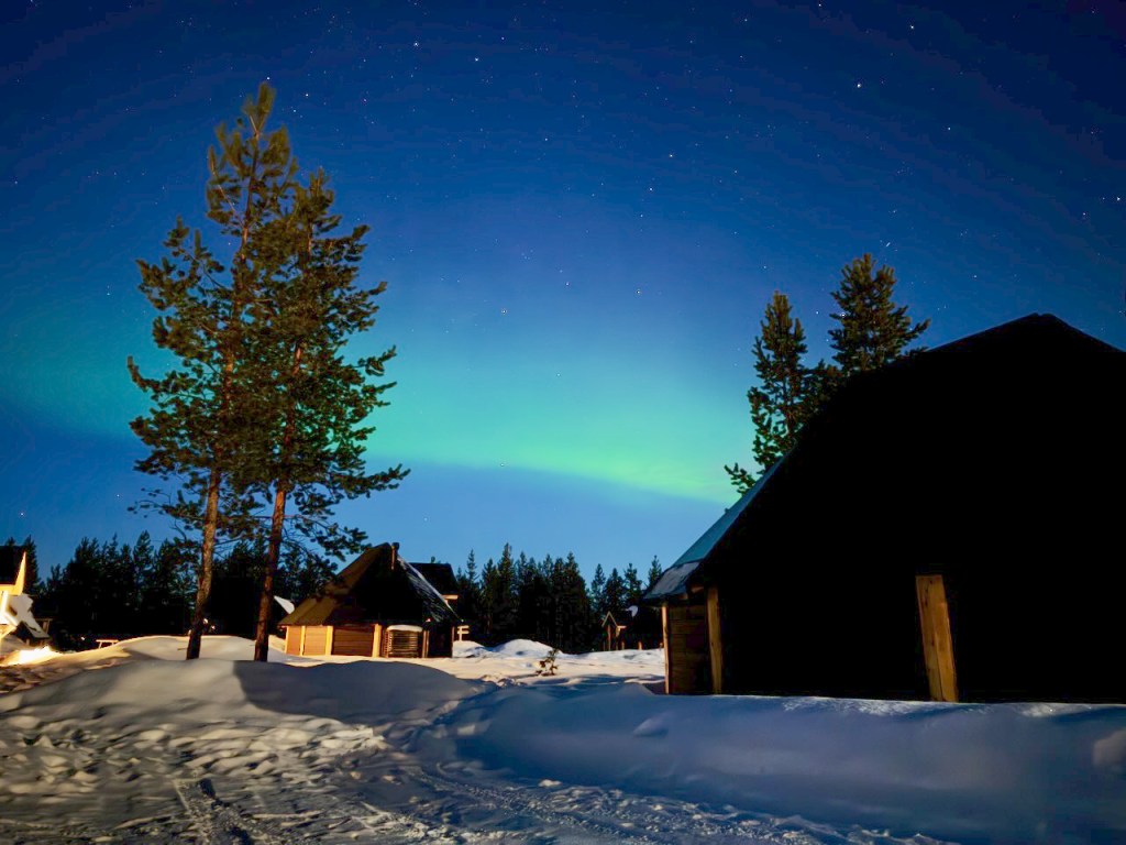 Night sky over a snowy landscape with cabins and pine trees, displaying a faint aurora borealis.