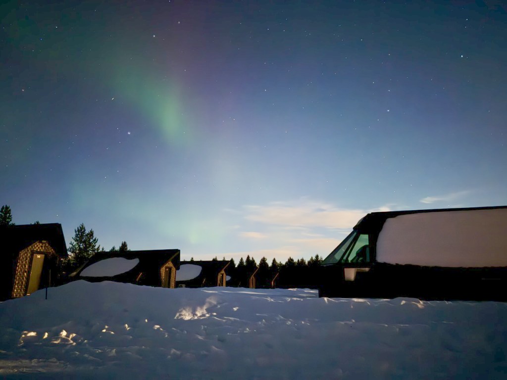 A night view of a snow-covered landscape with individual cabins under a starry sky, showcasing the northern lights in the background.