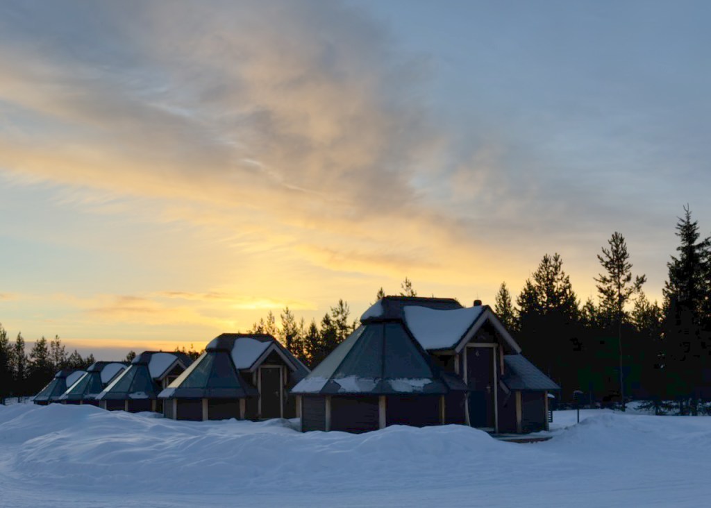 A row of 'Aurora Cabins' with snow-covered roofs, surrounded by a snowy landscape and evergreen trees, under a colorful sunset sky.