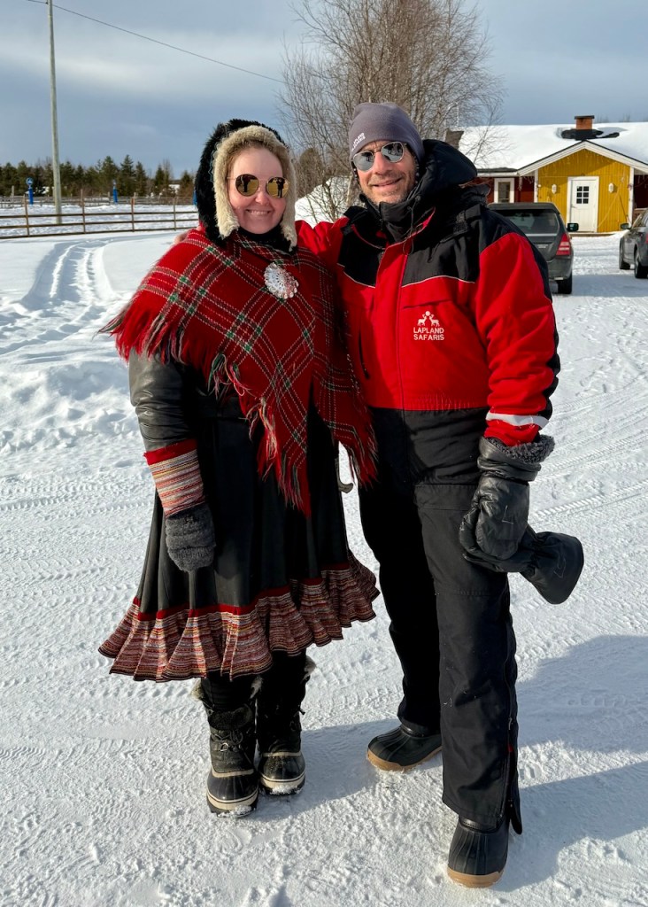 Two people standing together in a snowy landscape, wearing winter clothing. The woman on the left is dressed in a traditional red plaid blanket over a black dress, while the man on the right is wearing a red and black snowmobiling suit.