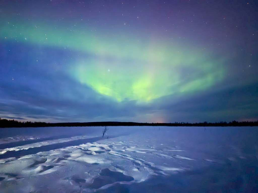 A stunning view of the Northern Lights illuminating the night sky over a snowy landscape, with a hint of purple and green hues reflected above a flat expanse of snow.