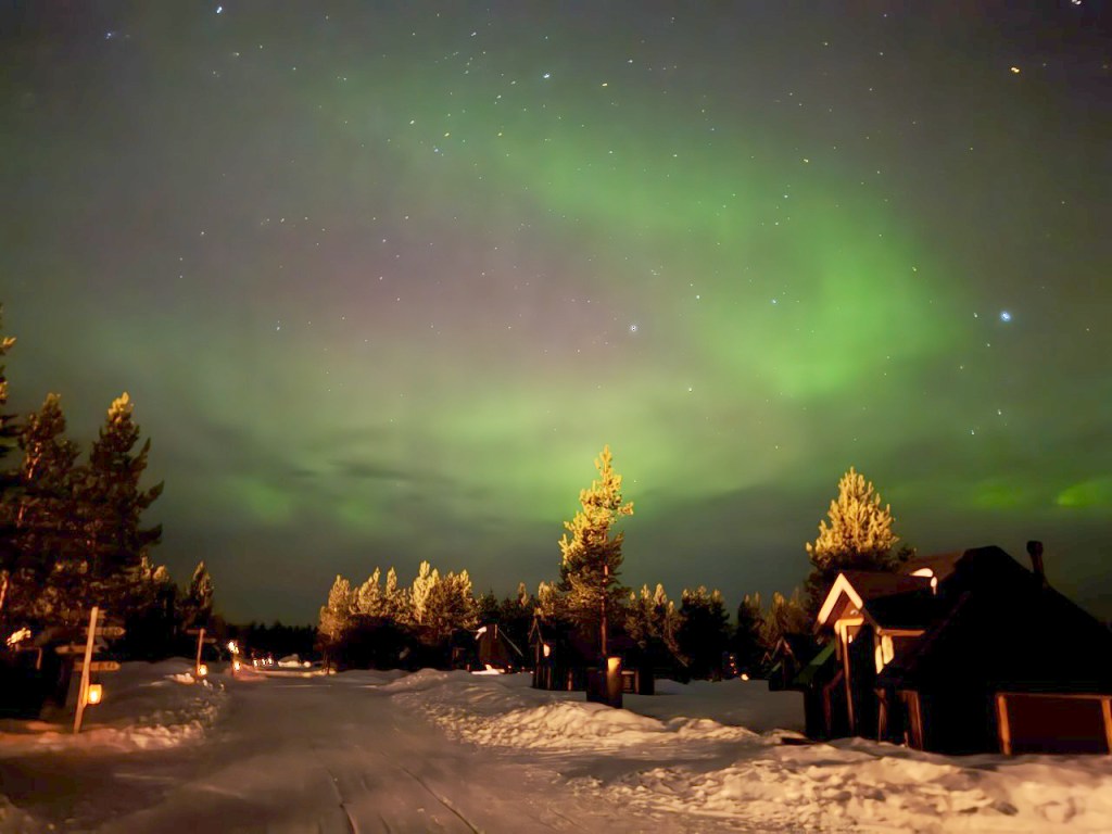 Northern lights illuminating the sky over a snowy landscape in Finnish Lapland, with cabins visible in the foreground.