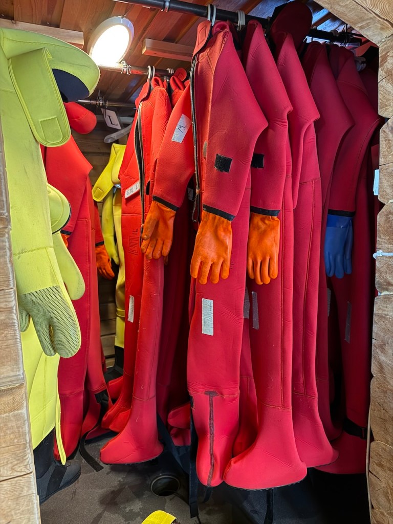 A variety of insulated wetsuits hanging in a storage area, including red and yellow suits, with orange and blue gloves visible.