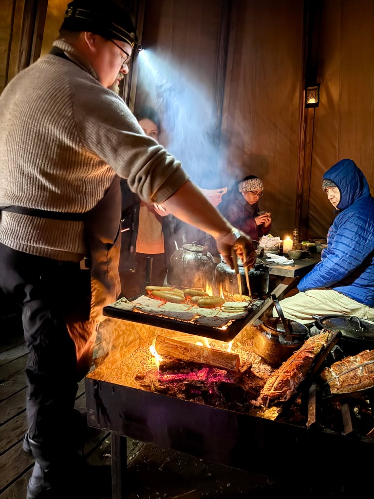A chef cooking over an open fire in a lavvu, with guests seated at a table nearby, in a cozy atmosphere with smoke rising and candles lit.