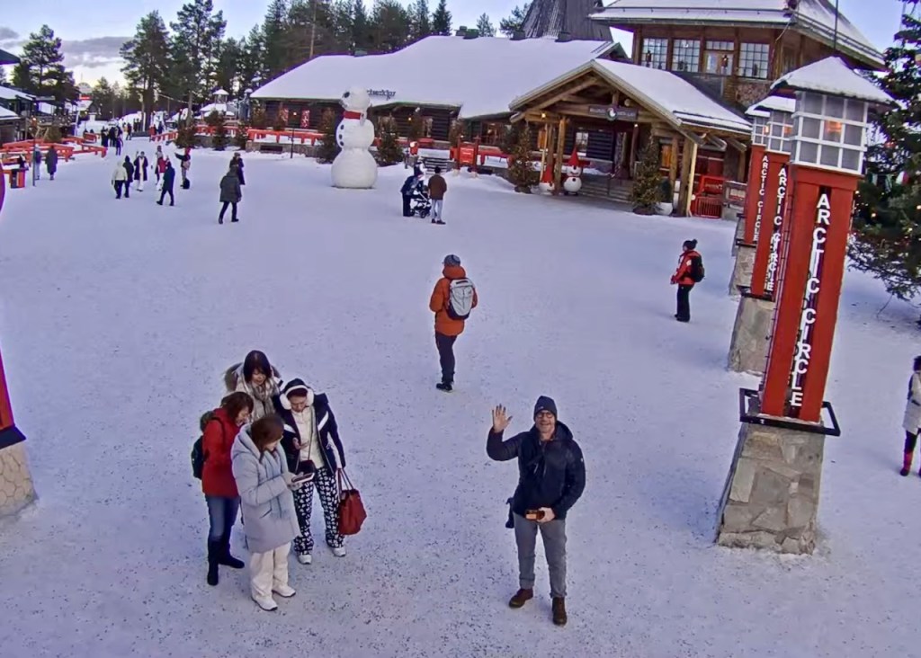 A snowy outdoor scene featuring a bustling area with people, a large snowman in the background, and signs indicating the Arctic Circle. Several visitors are gathered, with some looking at a device, while others walk around the snowy terrain.