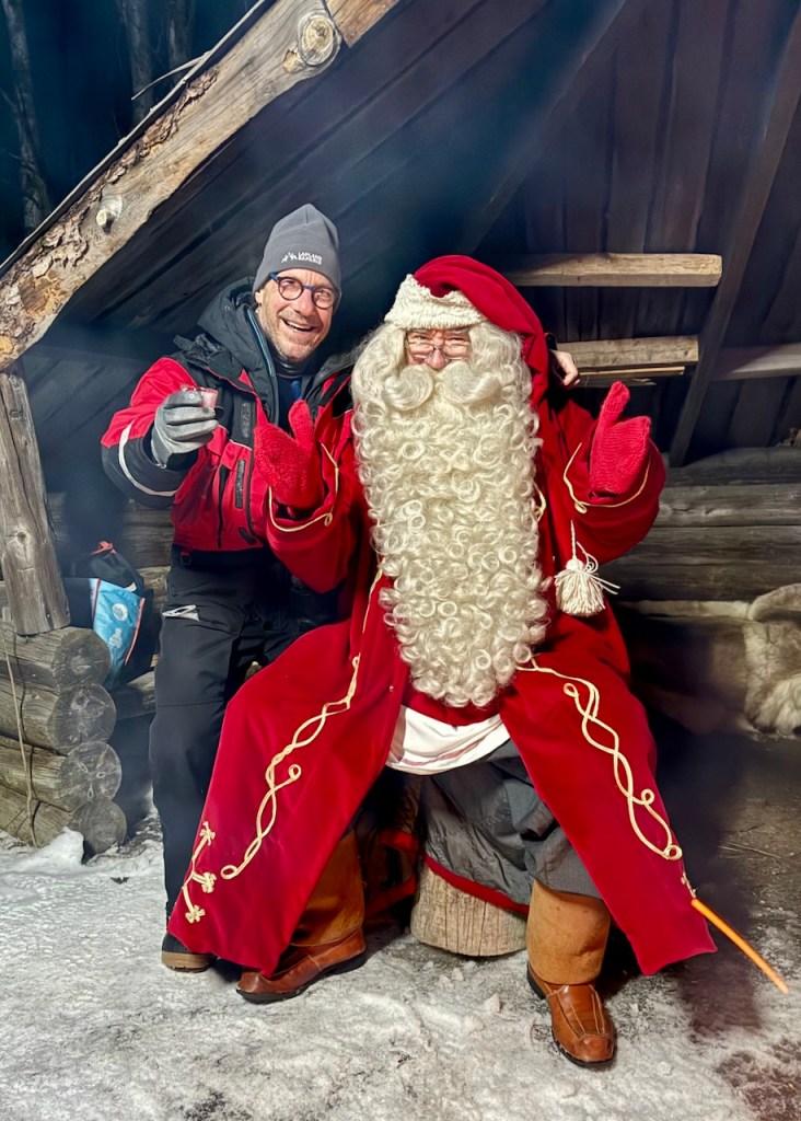 Two men posing together, one dressed in casual outdoor clothing and the other in a traditional Santa Claus outfit with a long white beard, sitting on a log in a wooden cabin setting.
