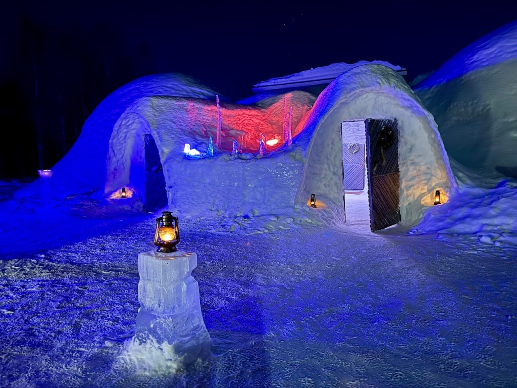An illuminated igloo restaurant at night, with colorful lights and lanterns, surrounded by snow.