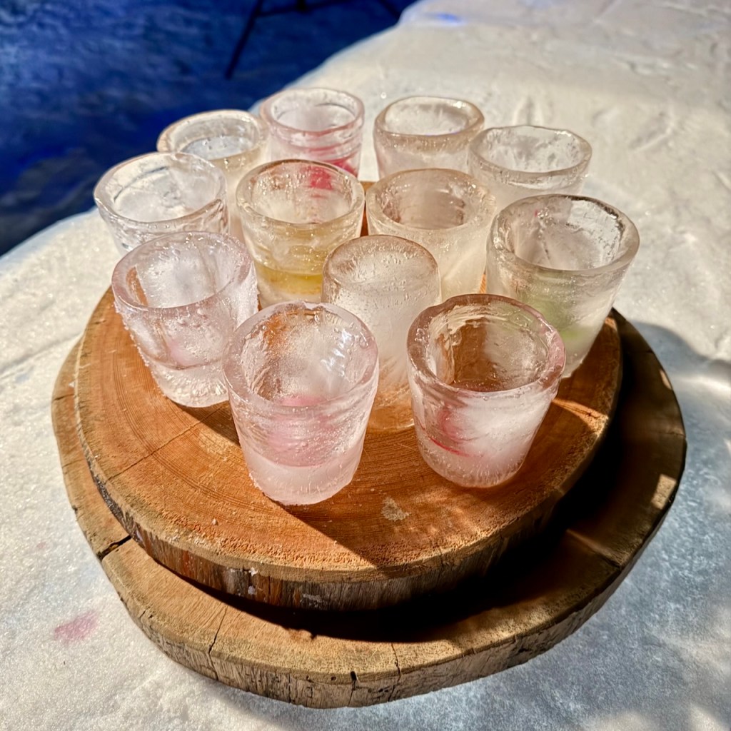 A wooden platter holding several shot glasses made of clear ice, with some appearing colored, arranged on a snowy surface.