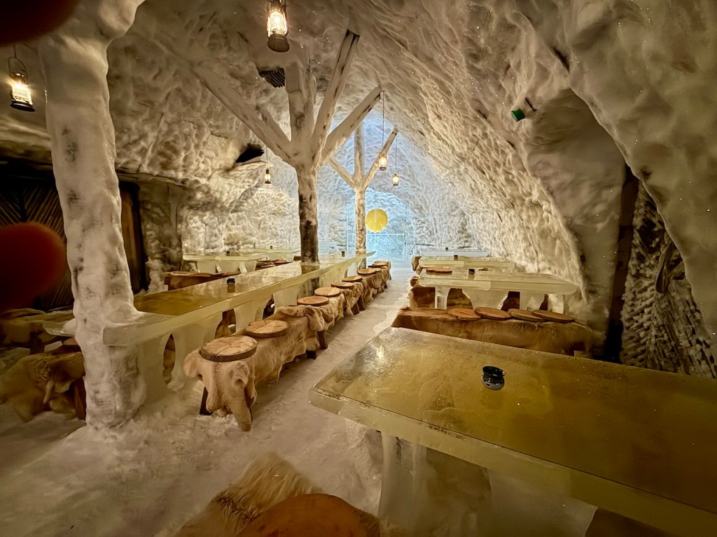 Interior of a unique igloo restaurant featuring icy walls, wooden furniture, and a warm ambiance with lanterns hanging from the ceiling.