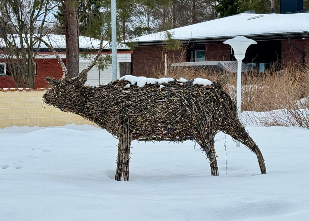 A decorative reindeer sculpture made of branches and twigs, partially covered with snow, situated in a snowy landscape near a suburban house.