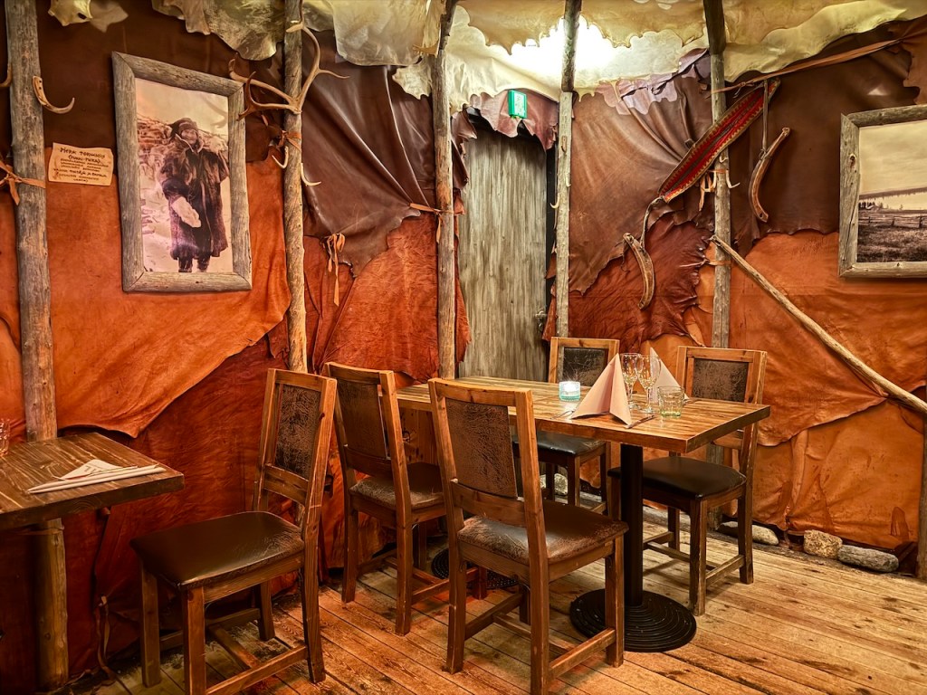 Interior of a rustic hobbit-style restaurant with wooden tables and chairs, decorated with animal skins and historical photos on the walls.