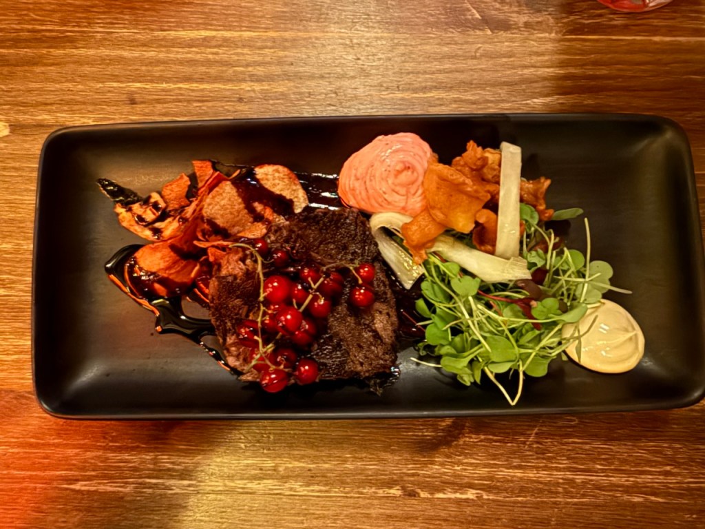 A beautifully plated dish featuring grilled reindeer steak garnished with red currants, creamy pink sauce, assorted vegetables, and microgreens on a black rectangular plate.
