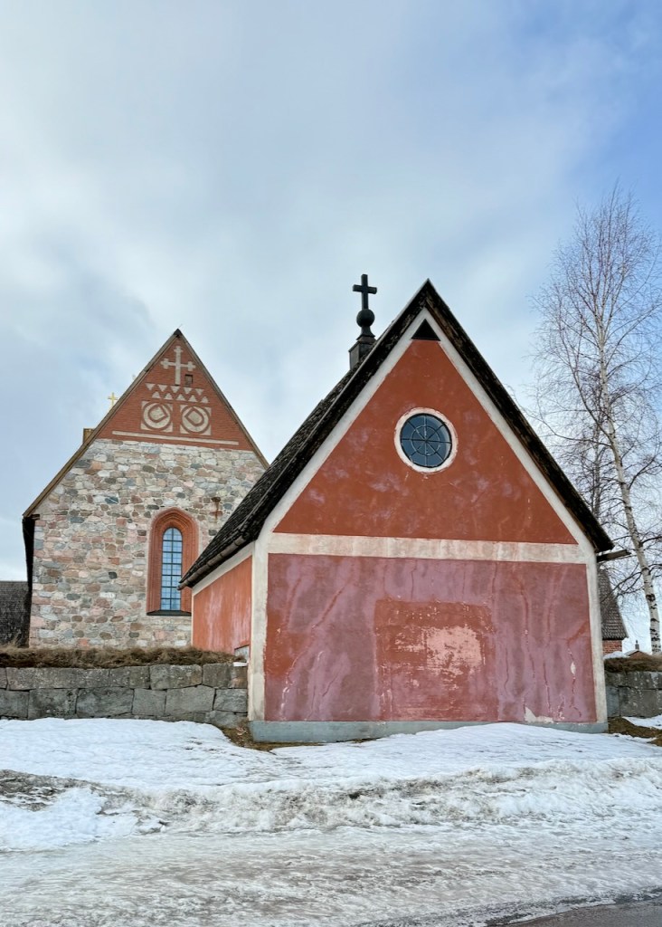Historic church buildings with distinct architectural styles, including a stone structure with a tall arched window and a smaller red building nearby, surrounded by snow and trees under a cloudy sky.