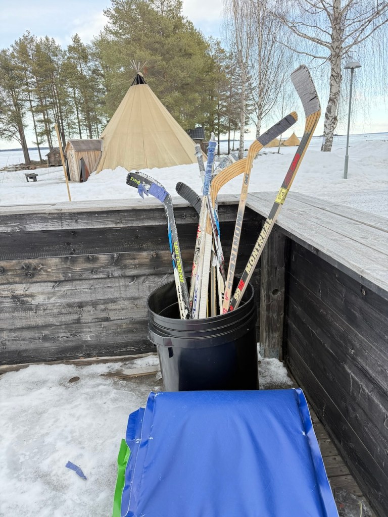 A collection of ice hockey sticks placed in a trash bin with a blue sled nearby, snowy ground, and a traditional Sami lavvo structure in the background.