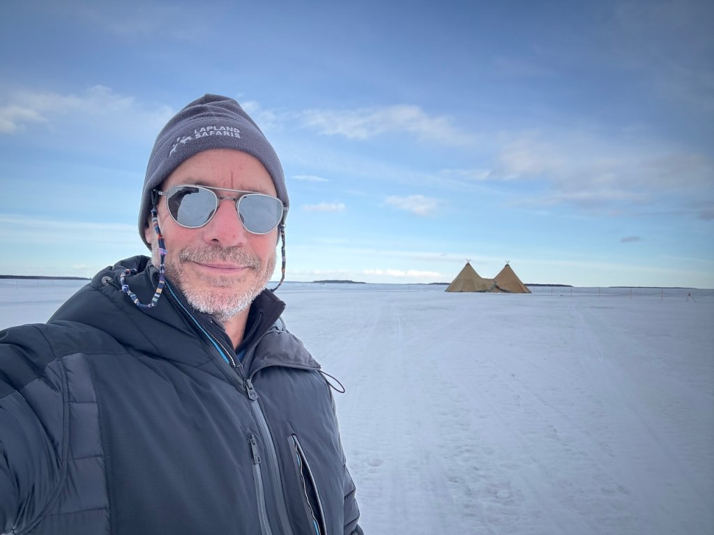A person wearing sunglasses and a winter jacket stands on a snowy landscape, with two tipis visible in the background under a clear blue sky.