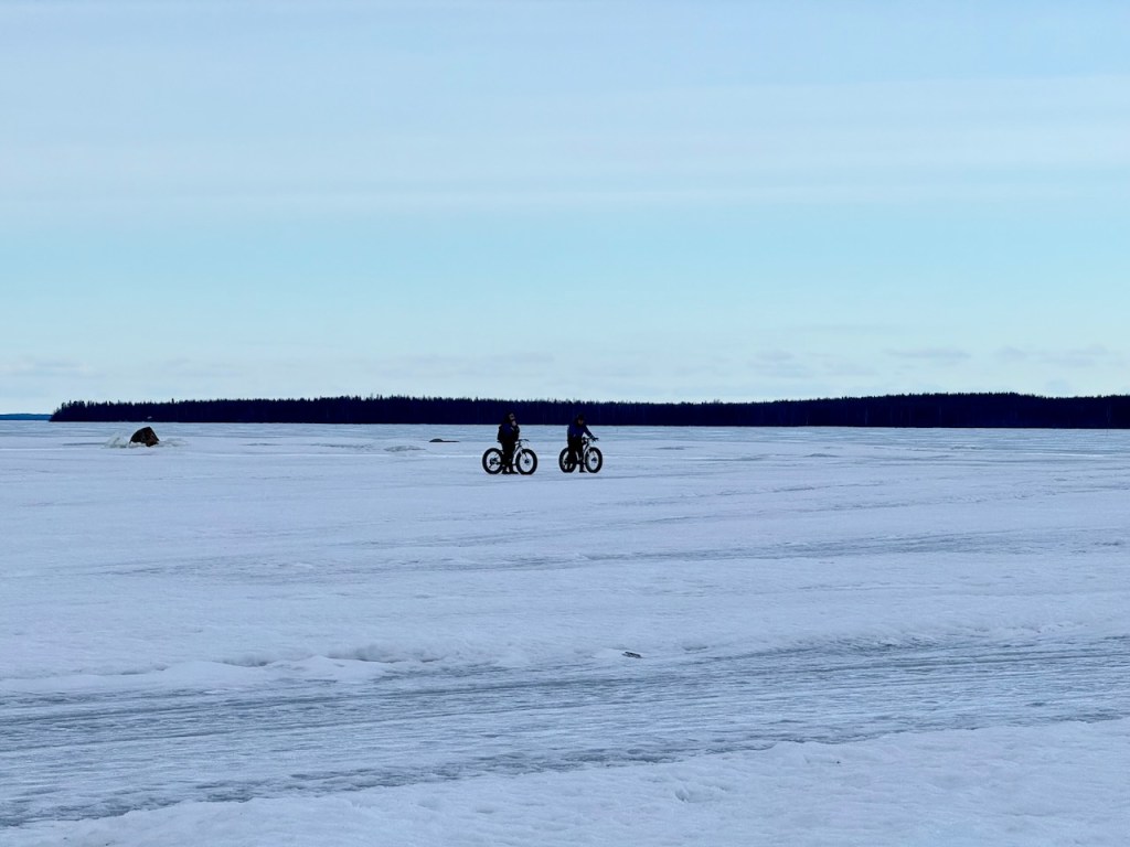 Two individuals riding fat bikes across a frozen surface on the Baltic Sea, with a snowy landscape in the background.