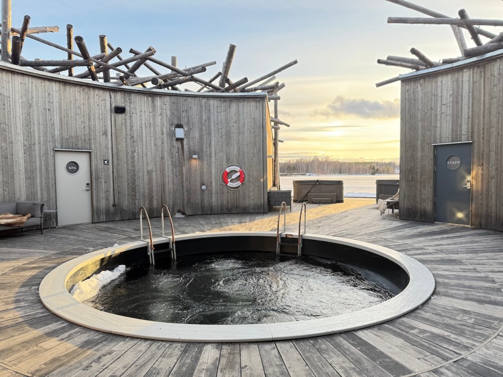A circular hot tub with water swirling inside, surrounded by wooden decking and buildings with rustic wooden exteriors. The sky shows a soft sunset with clouds, creating a serene atmosphere. A 'SPA' sign is visible on one of the doors.