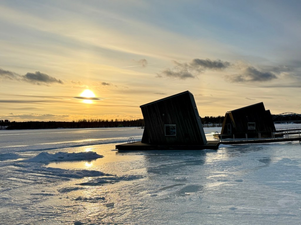 A scenic view of modern, angled wooden structures on a frozen river, surrounded by a serene winter landscape and a sunset in the background.