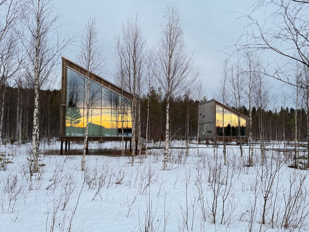 Modern architectural structures with large glass windows reflecting the sunset, surrounded by snow-covered ground and birch trees.