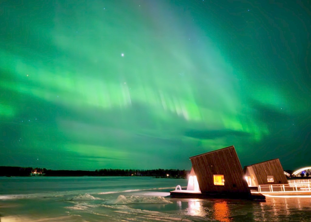 The Northern Lights illuminate the night sky with vibrant green hues above a frozen lake, featuring floating wooden structures in the foreground.