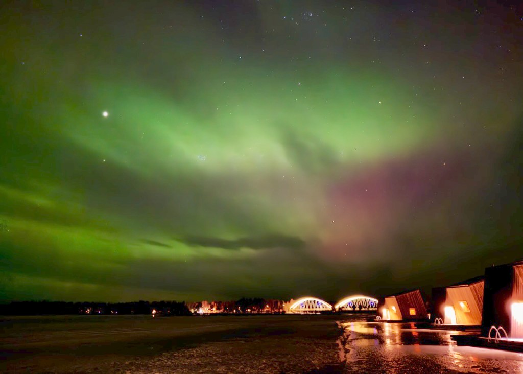 A stunning display of the Northern Lights over a serene landscape, with a river in the foreground and modern buildings softly illuminated by their warm lights.