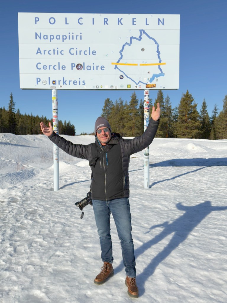 A man standing excitedly in front of a sign indicating the Arctic Circle, surrounded by snow-covered landscape and pine trees.