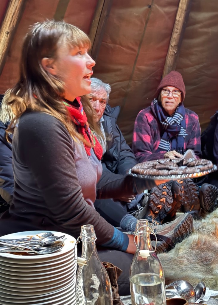 A Sami woman sharing food with guests while sitting in a lavvo, surrounded by attendees and traditional items.