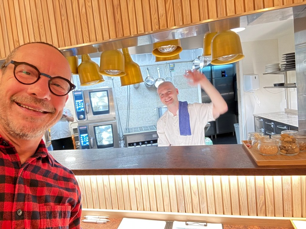 A smiling man in glasses posing for a selfie with a chef in the background waving. The kitchen is visible, featuring cooking equipment and utensils, with yellow hanging cups above.