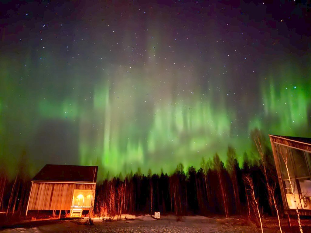 A stunning display of the Northern Lights against a night sky, with a silhouetted cabin and trees in the foreground.