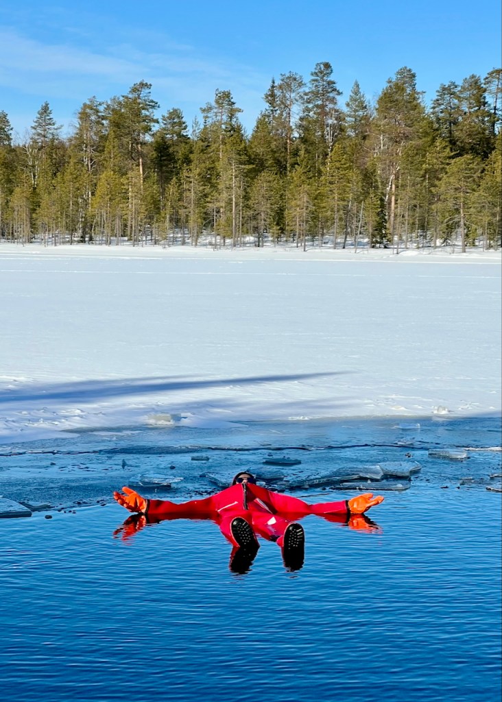 A person floating in a red suit on a frozen lake, with snowy banks and a backdrop of evergreen trees under a clear blue sky.