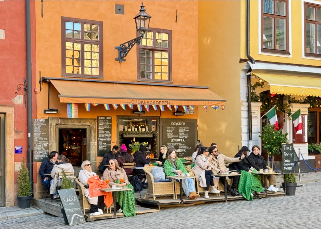 Outdoor café scene in Stockholm's Gamla Stan, featuring patrons enjoying drinks and food while seated at tables with green blankets, surrounded by colorful buildings.