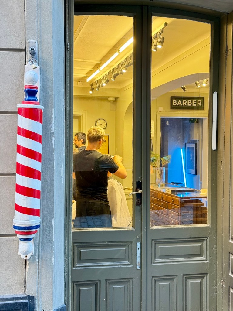 A barber shop interior view showing a barber cutting a client's hair, with a striped barber pole visible at the entrance.